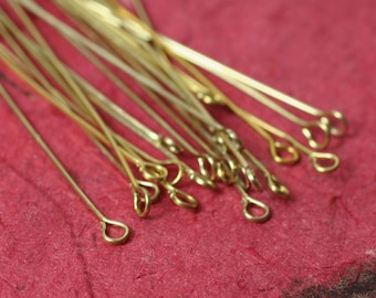 May include: A collection of gold-colored eye pins, each with a looped end, scattered on a textured red surface. The pins are thin and straight, ideal for jewelry making and crafting projects. The eye pins vary in length.