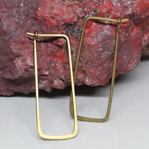 May include: A pair of gold-colored rectangular hoop earrings. The earrings have a simple, minimalist design with rounded corners and a hook closure. The earrings are displayed against a red and brown textured background.