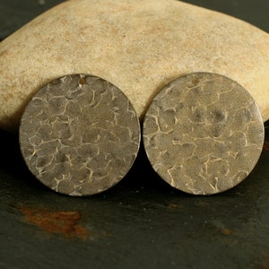 以下が含まれることがあります： ハンマー加工が施された、質感のある丸い金属製ペンダント2つ。各ペンダントには、紐を通すための小さな穴があります。ペンダントはくすんだブロンズ色で、暗い表面に表示されています。