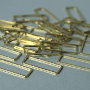 May include: A collection of small, rectangular brass metal frames. The frames are open in the centre and have a slightly textured surface. They are scattered across a light grey surface.