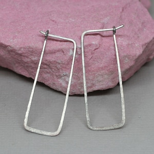 May include: A pair of silver rectangular hoop earrings. The earrings are displayed on a pink stone surface against a gray background. The earrings have a simple, minimalist design.