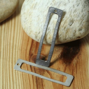 May include: Two silver metal rectangular tag shapes with a hole at the top. The tag on the left is smaller and has a smooth surface. The tag on the right is larger and has a textured surface.