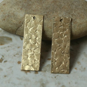 May include: Two rectangular, textured brass pendants with a hammered finish. Each pendant has a small hole at the top for stringing. The pendants are a warm, golden color.