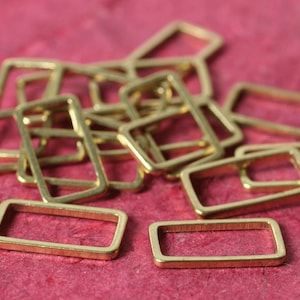 May include: A selection of small, gold-coloured rectangular metal findings. The open rectangles with rounded corners are scattered on a pinkish-red surface. These findings are likely used in jewellery making or other crafts.