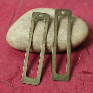 Puede incluir: Dos colgantes rectangulares de color latón antiguo con un recorte rectangular y un pequeño agujero en la parte superior para ensartar. Los colgantes se muestran sobre una piedra beige sobre un fondo rojo.
