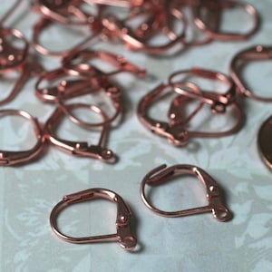 May include: Close-up of several rose gold leverback earring findings. The findings are made of metal and have a curved design with a hinged lever closure. A penny is visible in the background, providing a size reference. These findings are ideal for jewelry making.