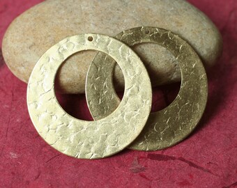 May include: Two gold-colored, hammered-texture hoop earrings. The earrings are circular with a hole at the top for hanging. The earrings are displayed on a red surface with a tan rock in the background.