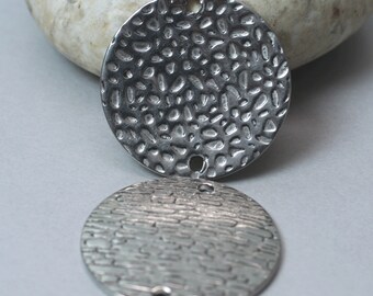 May include: Two silver-toned, textured metal pendants. The top pendant has a hammered, irregular pattern, while the bottom pendant has a linear, textured design. Each pendant has a small hole at the top for stringing. These pendants are suitable for jewelry making.