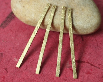 May include: Four textured, gold-colored metal bar pendants with small holes at the top. The bars are arranged on a red surface with a beige stone in the background. The pendants have a hammered texture.