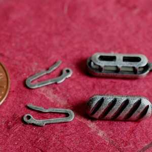 May include: Close-up of jewelry findings, including two silver-toned clasps and two decorative barrel-shaped clasps. One barrel clasp has a ribbed design. The items are on a red surface, with a US penny for scale.