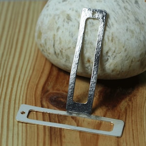 May include: Two silver metal rectangular pendant charms with a textured surface. The larger charm has a rectangular opening in the center. The smaller charm has a hole at the top.