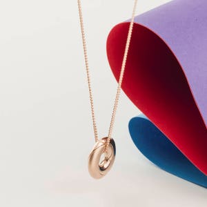 May include: A gold necklace with a simple, round pendant. The necklace is hanging against a backdrop of colorful paper.