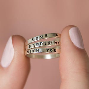 May include: Stackable silver rings with the words "LOVE & HAPPINESS WITH YOU" stamped on them. The rings are held between two fingers against a soft pink background. The rings are a simple, elegant design.