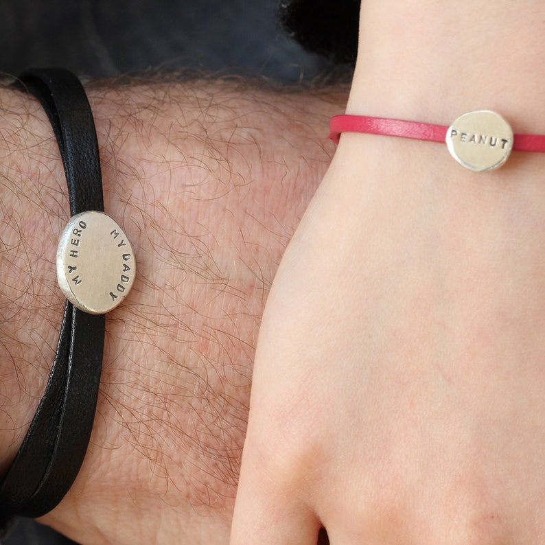 K&ouml;nnte beinhalten: Zwei Lederarmb&auml;nder mit silbernen Anh&auml;ngern. Das Armband links hat einen Anh&auml;nger mit der Aufschrift "My Daddy My Hero". Das Armband rechts hat einen Anh&auml;nger mit der Aufschrift "Peanut".