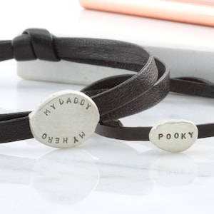 K&ouml;nnte beinhalten: Zwei schwarze Lederarmb&auml;nder mit silbernen ovalen Anh&auml;ngern. Ein Anh&auml;nger sagt "MY DADDY MY HERO" und der andere sagt "POOKY".