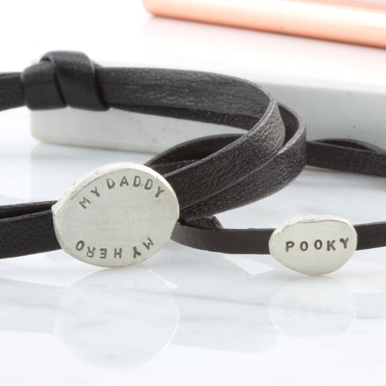K&ouml;nnte beinhalten: Zwei schwarze Lederarmb&auml;nder mit silbernen ovalen Anh&auml;ngern. Ein Anh&auml;nger sagt "MY DADDY MY HERO" und der andere sagt "POOKY".