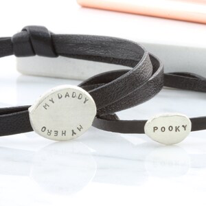 K&ouml;nnte beinhalten: Zwei schwarze Lederarmb&auml;nder mit silbernen ovalen Anh&auml;ngern. Der gr&ouml;&szlig;ere Anh&auml;nger tr&auml;gt die Aufschrift "MY DADDY MY HERO" und der kleinere Anh&auml;nger die Aufschrift "POOKY".