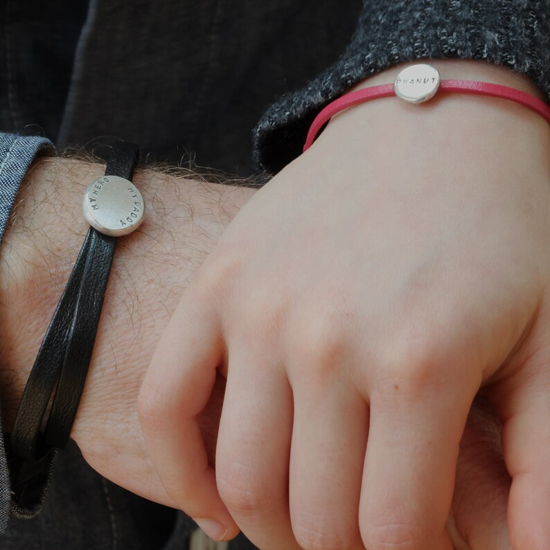 K&ouml;nnte beinhalten: Zwei Lederarmb&auml;nder mit runden Silberanh&auml;ngern. Das Armband links hat ein schwarzes Lederband und der Anh&auml;nger sagt "TODAY YOU ARE MY HERO". Das Armband rechts hat ein pinkfarbenes Lederband und der Anh&auml;nger sagt "DONUT".