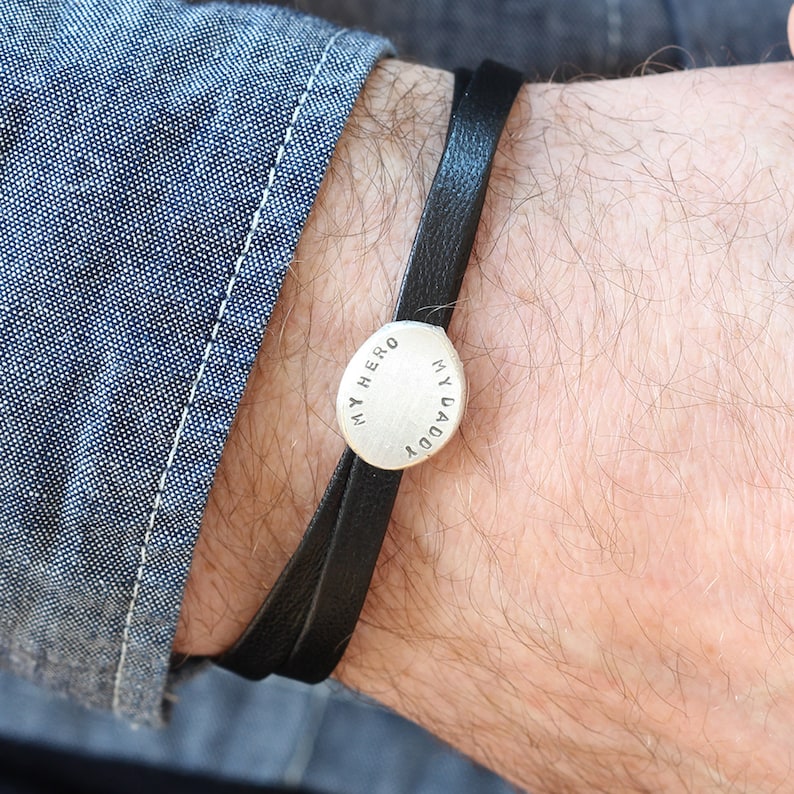 K&ouml;nnte beinhalten: Ein schwarzes Lederarmband mit einem silbernen Anh&auml;nger, auf dem "Mein Held, mein Papa" steht.