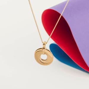 May include: A gold necklace with a circular pendant. The pendant is made of two overlapping circles, creating a unique design. The necklace is displayed against a colorful background.