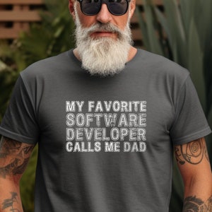 以下が含まれることがあります： 白文字で「私のお気に入りのソフトウェア開発者は私をパパと呼ぶ」と書かれた濃いグレーのTシャツ