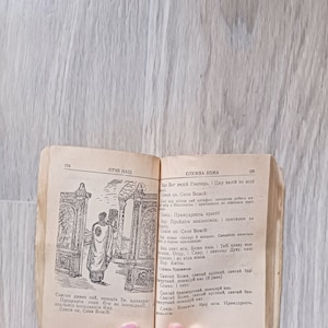May include: An open book with a black and white illustration of a person in a robe standing in a doorway. The book has the text "Отче наш" on the left page and "СЛУЖБА БОЖА" on the right page. The text on the right page also includes "Яво Бог велій Господь, і Цар велій по всей" and "Спаси ни, Сине Божій".