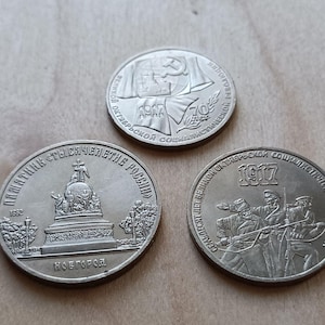 May include: Three circular silver-toned commemorative coins. The top coin features a hammer and sickle with the text 1917 and 70 ЛЕТ. The bottom left coin depicts a monument with the text ПАМЯТНИК ТЫСЯЧЕЛЕТИЕ РОССИИ. The bottom right coin shows figures with 1917.