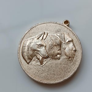 May include: A round, gold-colored pendant featuring embossed images of three different dog breeds. The pendant has a small loop at the top for hanging. The dogs are in profile, with detailed fur and facial features.