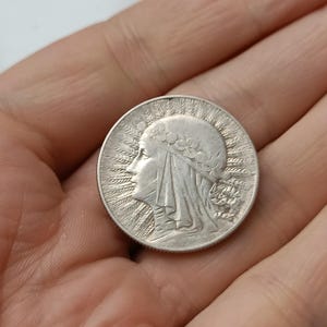 Puede incluir: Una moneda de plata que presenta el perfil de una mujer con un tocado drapeado y adornos florales. La moneda tiene un diseño detallado con líneas radiales alrededor del borde. La moneda se sostiene en una mano.