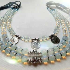 Puede incluir: Collar de varias hebras con cuentas opalinas y detalles plateados. El collar tiene tres hebras de cuentas redondas, elementos decorativos plateados y colgantes en forma de moneda. Cintas de terciopelo azul oscuro sirven como lazos.