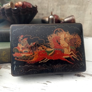 May include: A rectangular black lacquer box featuring a hand-painted scene of a figure in a red coat riding in a sleigh pulled by three horses. The artwork includes gold and red details, depicting a dynamic winter scene with swirling lines.