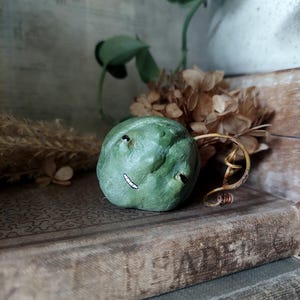 Könnte beinhalten: Eine kleine, runde, grüne Skulptur, die einem Kürbis ähnelt, mit strukturierter Oberfläche, zwei dunklen, mandelförmigen Augen und einem dünnen, weißen, handgemalten Mund. Ein dünner, brauner, gewundener Stiel ragt rechts aus dem Objekt.