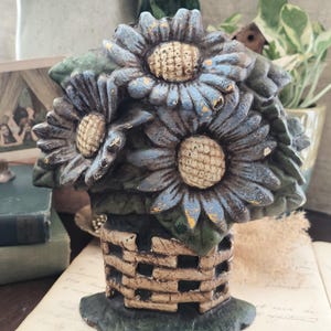 May include: A cast iron doorstop shaped like a bouquet of three blue sunflowers with textured cream centers, set in a tan woven basket base. The metal surface features a distressed, weathered finish with visible green leaves and dark paint accents.