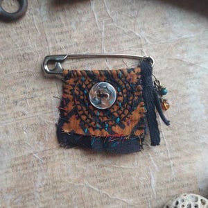 May include: A safety pin brooch featuring a small square of orange and black patterned fabric with frayed edges. The fabric is embellished with dark stitching, a clear plastic button in the center, and a black fabric strip with small dangling beads.