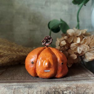 Könnte beinhalten: Eine kleine, orangefarbene, handgefertigte Kürbisfigur mit strukturierter Oberfläche, einem dunkelbraunen, gedrehten Stiel, zwei winzigen schwarzen Perlenaugen, einer markanten zentralen Nase und einem kleinen weißen Mund mit Zähnen.