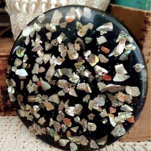 May include: A circular black coaster featuring an embedded mosaic of iridescent shell fragments in various shapes and sizes. The center of the coaster displays the gold-colored text THE OZERKS, surrounded by the scattered, shimmering shell pieces.