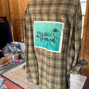 May include: A green and brown plaid flannel shirt with a turquoise blue patch on the back. The patch has a dandelion design and the text "BRILLIANTLY Untamed".