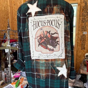 May include: Green and black plaid flannel shirt with a "Hocus Pocus" graphic on the back. The graphic features the movie's title and characters. Two white star-shaped patches are sewn on the shirt.