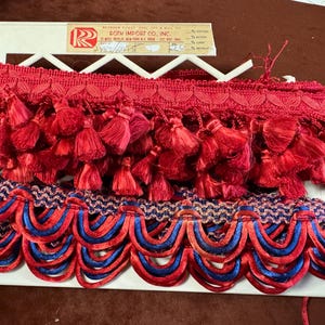May include: Red tassel fringe trim with a scalloped blue and red design. The trim is displayed on a white plastic holder. The top layer features a row of red tassels, and the bottom layer has a wavy pattern of blue and red.