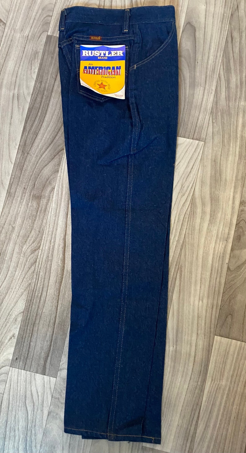 May include: A pair of dark blue denim jeans with a yellow and red label that reads "Rustler Brand An American Tradition" in the back pocket.