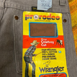 Puede incluir: Una etiqueta amarilla y roja con el texto "ProRodeo Endorsed Jeans" y un logotipo de Wrangler. La etiqueta presenta un vaquero de dibujos animados tirando de una cuerda con un toro. El texto "Boys Cowboy Cut" est&aacute; impreso en la etiqueta. La etiqueta tambi&eacute;n incluye el texto "Our Original 100% Cotton 14 3/4 Ounce Pre-Shrunk Denim Jeans".