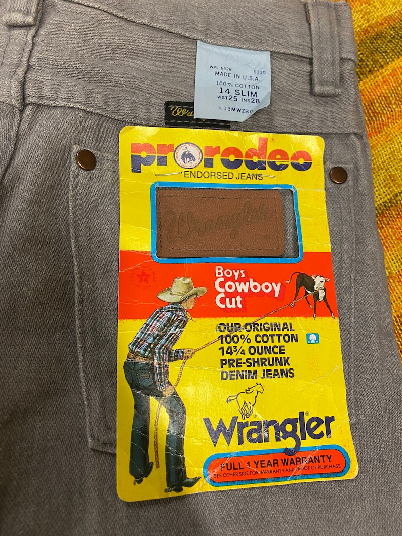 Puede incluir: Una etiqueta amarilla y roja con el texto "ProRodeo Endorsed Jeans" y un logotipo de Wrangler. La etiqueta presenta un vaquero de dibujos animados tirando de una cuerda con un toro. El texto "Boys Cowboy Cut" est&aacute; impreso en la etiqueta. La etiqueta tambi&eacute;n incluye el texto "Our Original 100% Cotton 14 3/4 Ounce Pre-Shrunk Denim Jeans".