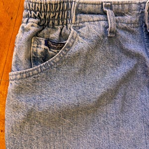May include: Close-up of light blue denim jeans with an elastic waistband and a front pocket. The pocket features a small, dark blue Wrangler label. The jeans have a faded, worn appearance.