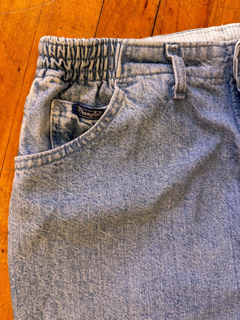 May include: Close-up of light blue denim jeans with an elastic waistband and a front pocket. The pocket features a small, dark blue Wrangler label. The jeans have a faded, worn appearance.