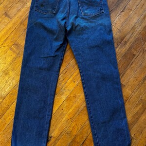 May include: Dark blue denim jeans with a classic five-pocket design. The jeans feature a brown leather patch with the brand name on the back pocket. The jeans are a straight-leg style.