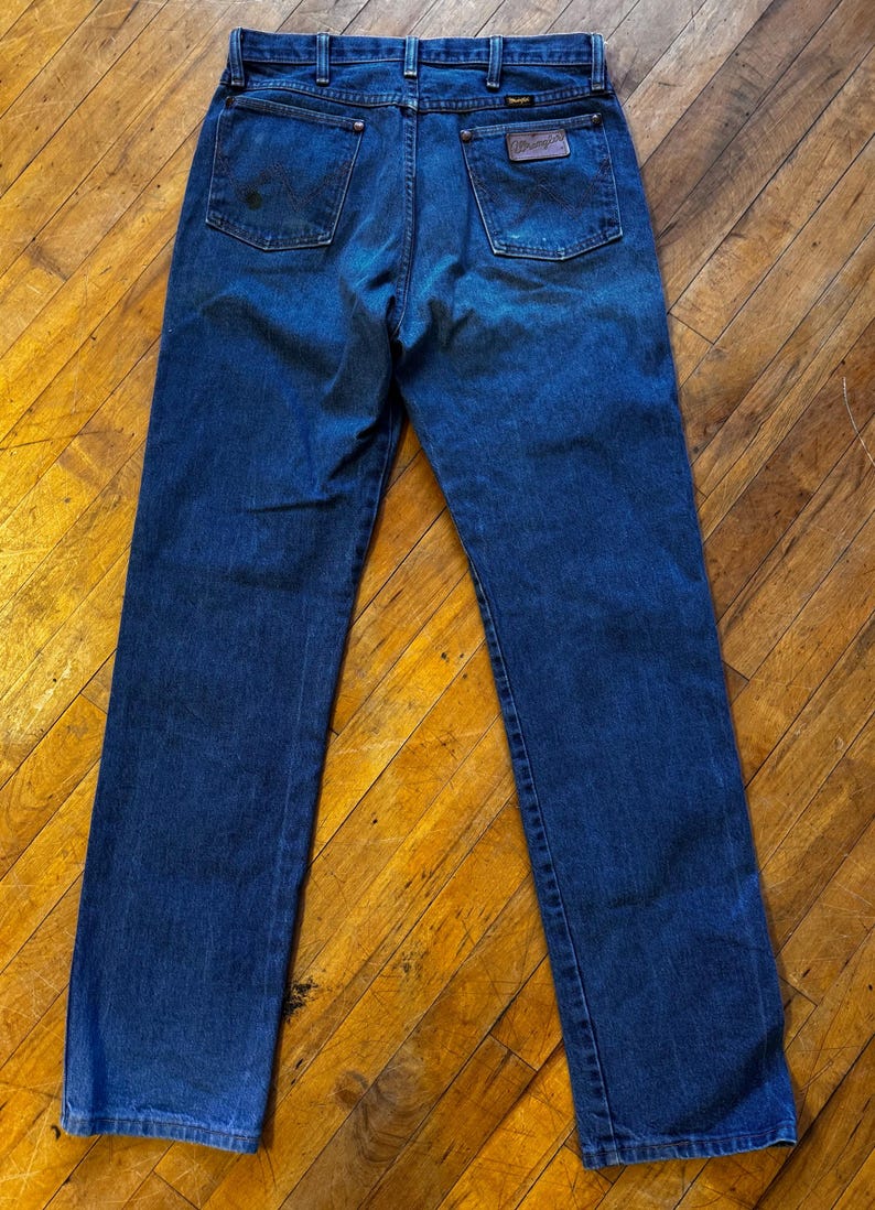 May include: Dark blue denim jeans with a classic five-pocket design. The jeans feature a brown leather patch with the brand name on the back pocket. The jeans are a straight-leg style.