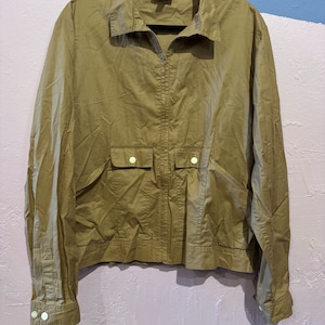 May include: A khaki green zip-up jacket with a collared neckline and two large pockets on the front. The jacket has white buttons on the pockets and cuffs.
