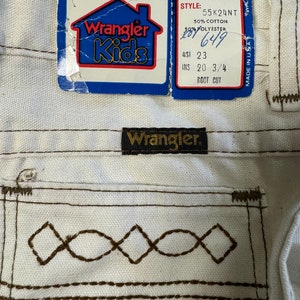 K&ouml;nnte beinhalten: Wei&szlig;e Wrangler Kids Jeans mit braunen N&auml;hten und einem Etikett mit der Aufschrift "Wrangler Kids" und "Full 1 Year Warranty". Das Etikett enth&auml;lt auch die Gr&ouml;&szlig;e, den Stil und die Stoffzusammensetzung. Die Jeans sind in den USA hergestellt.