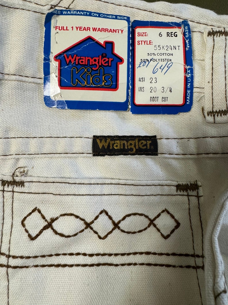K&ouml;nnte beinhalten: Wei&szlig;e Wrangler Kids Jeans mit braunen N&auml;hten und einem Etikett mit der Aufschrift "Wrangler Kids" und "Full 1 Year Warranty". Das Etikett enth&auml;lt auch die Gr&ouml;&szlig;e, den Stil und die Stoffzusammensetzung. Die Jeans sind in den USA hergestellt.