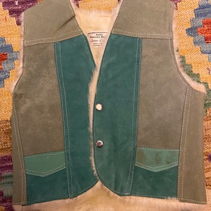 May include: A vintage suede vest with a sheepskin lining. The vest features a patchwork design in shades of green and teal, with white stitching and two silver buttons. The interior is lined with cream-colored sheepskin. The vest has a label that reads "Genuine Suede".